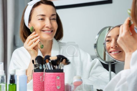 Foto de Retrato sonriente de una mujer adulta vestida con bata en el baño use rodillo facial para el cuidado de la piel frente al espejo - Imagen libre de derechos