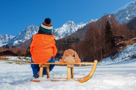 Téléchargez les photos : Petit garçon avec lapin en peluche dans la tenue d'hiver assis sur un traîneau en bois sur la haute montagne vue de derrière - en image libre de droit