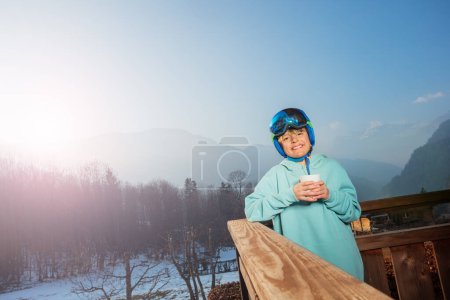 Foto de Sonriente niño esquiador adolescente de pie en la mañana en casco y sudadera en el balcón, listo para esquiar en las montañas nebulosas - Imagen libre de derechos
