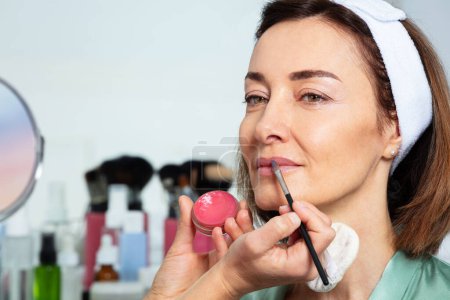 Téléchargez les photos : Portrait en gros plan d'une esthéticienne utilisant un pinceau pour maquiller les lèvres d'une femme d'âge moyen portant un bandeau assis devant un miroir - en image libre de droit