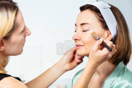Téléchargez les photos : Esthéticienne maquilleuse mettant de la crème de fond de teint de première qualité avec pinceau sur le visage d'une femme d'âge moyen portant un bandeau - en image libre de droit