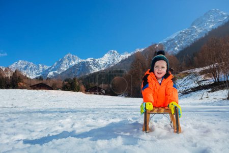 Foto de Niño sonriente en abrigo naranja de invierno y sombrero de lana yacía en el trineo deslizándose cuesta abajo sobre la nieve - Imagen libre de derechos