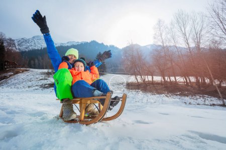 Téléchargez les photos : Deux garçons, frères en tenue d'hiver assis sur le traîneau descendant levant les mains souriantes et s'amusant en vacances - en image libre de droit