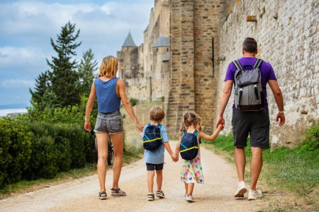 Foto de Joven familia turística con dos niños tienen vacaciones de verano en Francia visitando la fortaleza de Carcasona, caminando a lo largo de enormes paredes de piedra, disfrutando de monumento histórico, vista desde atrás - Imagen libre de derechos