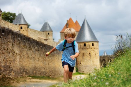 Foto de Pequeño aventurero niño escalada colina en el día de verano con Carcasona castillo fortificado francés en el fondo en Occitania, Francia - Imagen libre de derechos