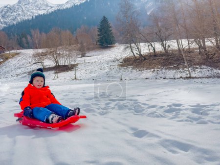 Photo for Little boy in winter outfit slide downhill on alpine slope in the mountains on red sledge - Royalty Free Image