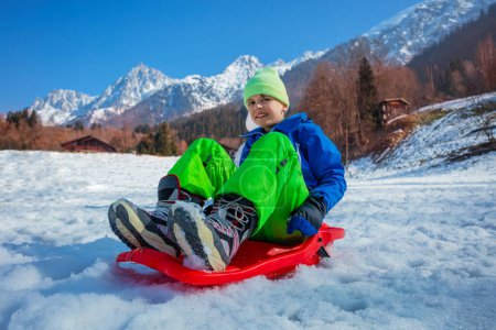 Photo for Young boy in winter outfit slide downhill on alpine slope in the mountains on red sledge - Royalty Free Image