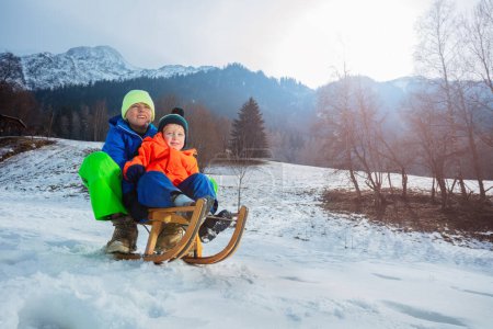 Téléchargez les photos : Deux garçons, frères en tenue d'hiver assis sur le traîneau descendant souriant et s'amusant en vacances - en image libre de droit