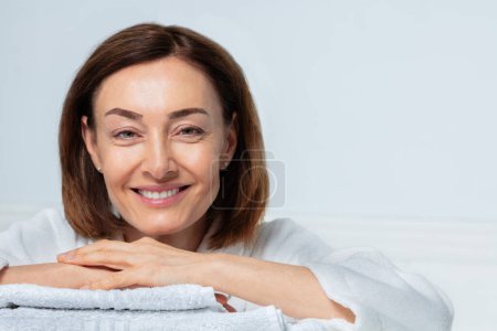 Foto de Retrato de belleza de cerca de una mujer de mediana edad, con una sonrisa confiada en la casa posando en la configuración del baño poniendo las manos en la toalla - Imagen libre de derechos