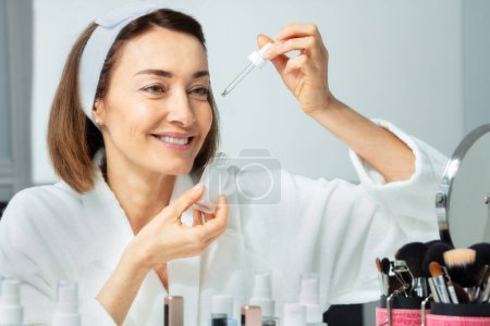 Photo for Close-up portrait of a beautiful middle aged woman putting facial serum with eyedropper on the face, wide smiling before make mirror - Royalty Free Image