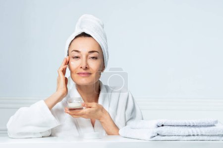 Téléchargez les photos : Photo de beauté d'une femme mettant de la crème de pot sur le visage avec turban serviette sur mur de salle de bain blanc - en image libre de droit