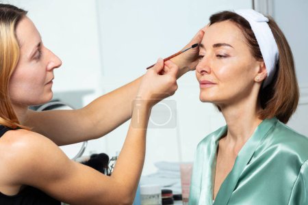 Foto de Esteticista aplicar rimel usando cepillo de cejas en la hermosa mujer de mediana edad antes de maquillaje espejo vista de perfil - Imagen libre de derechos