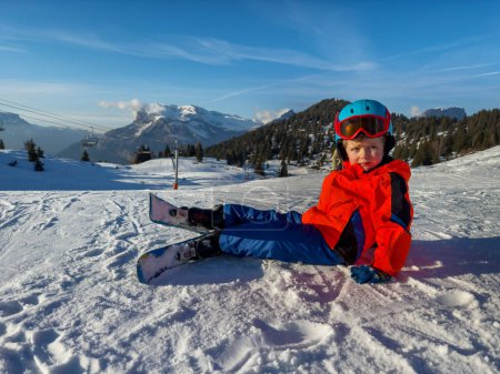 Foto de Niño esquiador sentado en la pista de esquí en casco y máscara tomar un descanso en las montañas soleadas francesas - Imagen libre de derechos