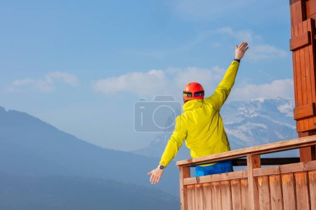 Photo for Happy man skier standing on balcony of French chalet in ski helmet and mask overlooking mountains, lifting hands - Royalty Free Image