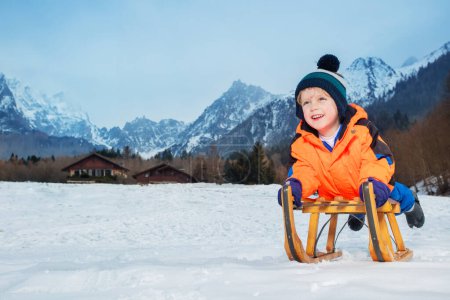 Téléchargez les photos : Petit garçon riant en manteau d'hiver orange et chapeau de laine allongé sur le traîneau glissant en descente sur la neige souriant - en image libre de droit