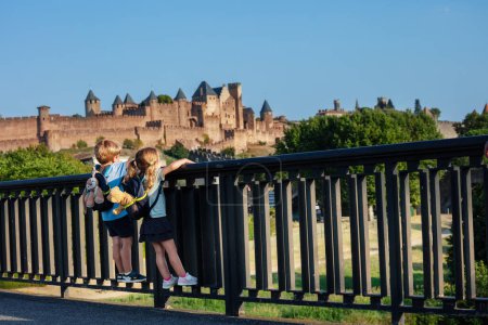 Téléchargez les photos : Deux petits enfants voyageurs avec de petits sacs à dos regardent le château de Carcassonne en été lumière du coucher du soleil depuis le pont suspendu à la balustrade, vue de derrière - en image libre de droit