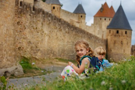 Foto de Gemelos, hermanos y hermanas exploran la histórica ciudad de Carcasona juntos sentados a lo largo de muros de piedra disfrutando de vacaciones de verano en el sur de Francia - Imagen libre de derechos