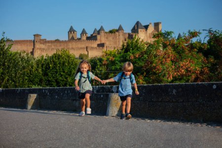 Téléchargez les photos : Des enfants heureux tenant la main courent sur un pont avec le château de Carcassonne en arrière-plan excités de commencer leurs vacances d'été dans le sud de la France - en image libre de droit