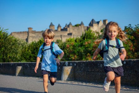 Foto de Niños felices mochileros corren con el castillo de Carcassonne en el fondo emocionados de comenzar sus aventuras de vacaciones de verano en Francia - Imagen libre de derechos