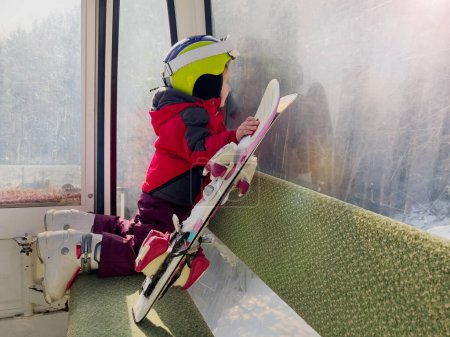 Foto de Joven esquiador con casco sentado en teleférico observando pistas de esquí sosteniendo esquís - Imagen libre de derechos
