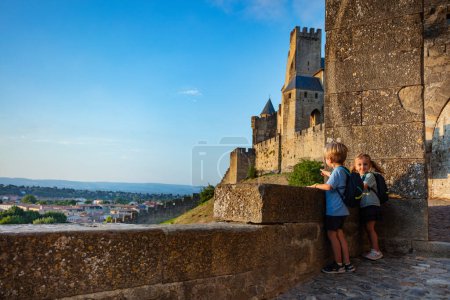 Foto de Dos pequeños viajeros sonrientes con pequeñas mochilas mirando a su alrededor desde la pared del castillo de Carcassonne en verano la luz del atardecer, viajando concepto de niños - Imagen libre de derechos