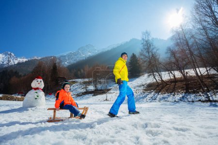 Foto de Vista del perfil de un padre tirando cuesta arriba de un niño en el trineo de madera sobre altas montañas y muñeco de nieve en invierno - Imagen libre de derechos