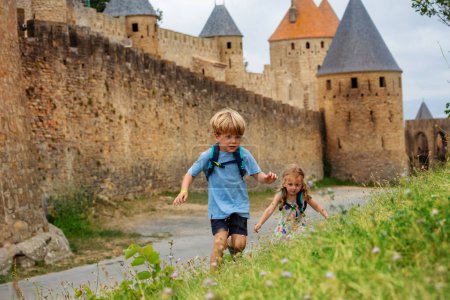 Foto de Dos pequeños turistas, niño y niña con mochilas explorando colinas de hierba de Carcasona fortaleza francesa en Occitania, Francia - Imagen libre de derechos