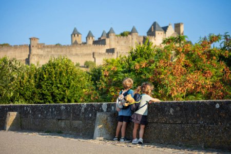 Téléchargez les photos : Deux petits enfants avec de petits sacs à dos regardent le château de Carcassonne au coucher du soleil d'été depuis le pont, vue de derrière, concept enfant voyageur - en image libre de droit