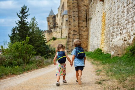 Foto de Los niños pequeños con mochilas de vacaciones hacen turismo corriendo a lo largo de enormes paredes de la ciudad fortificada francesa de Carcasona en Occitania, sur de Francia - Imagen libre de derechos