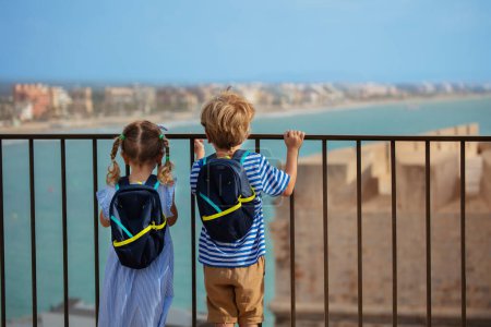 Foto de Dos niños pequeños, niño y niña visitando España disfrutan del mirador del castillo Peniscola en el soleado día de verano, miran a la bahía, ven desde atrás - Imagen libre de derechos