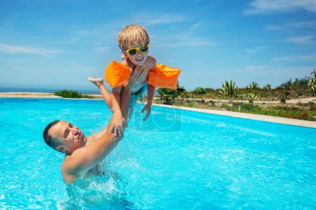 Téléchargez les photos : Dans la piscine adulte soulevant un enfant haut au-dessus de l'eau d'une manière ludique, garçon avec des lunettes de soleil portant des flotteurs sur leurs bras pour aider à nager - en image libre de droit
