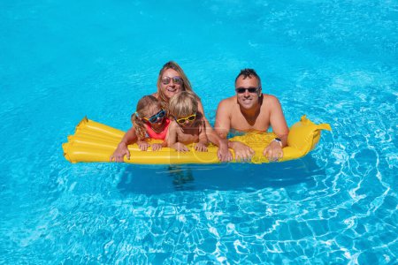 Téléchargez les photos : Une famille joyeuse avec des enfants prend plaisir à flotter tranquillement sur un matelas gonflable jaune dans les eaux calmes d'une piscine, flanqué d'un ciel lumineux et sans nuages - en image libre de droit