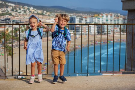 Foto de Dos lindos niños, chico, chica visitando España les encanta la vista de la costa, saltar y bailar en el punto de observación del castillo Peniscola en el soleado día de verano - Imagen libre de derechos