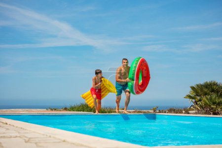 Téléchargez les photos : Un duo, papa et fils se prépare pour une baignade équipée de gonflables, à côté d'une piscine claire sur un ciel, fond de mer - en image libre de droit