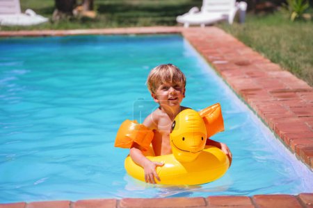 Foto de Un niño flota felizmente en una piscina sobre un anillo inflable de pato amarillo brillante, con brazaletes anaranjados durante su viaje de vacaciones de verano - Imagen libre de derechos