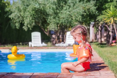 Foto de Un niño pequeño en un traje de baño naranja vibrante se sienta al lado de una piscina brillante, con exuberante vegetación en el fondo y un pato inflable flotando en el agua - Imagen libre de derechos
