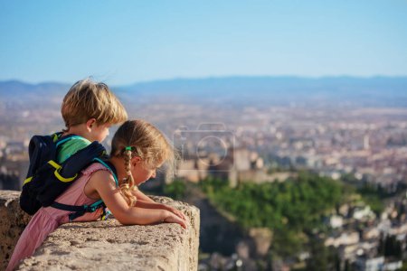 Foto de Pequeños viajeros en mirador admiran el antiguo palacio de la Alhambra en sus vacaciones de verano en Granada, España - Imagen libre de derechos