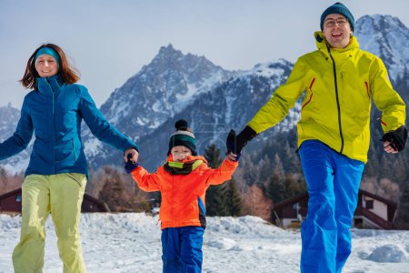 Foto de Padre y madre riendo corren tomados de la mano con su pequeño sol en el medio sobre montañas de nieve vistiendo traje de invierno - Imagen libre de derechos