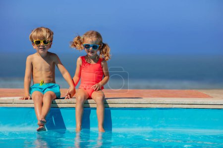 Foto de Jóvenes amigos, niños y niñas llevan gafas de sol sentados cómodamente al borde de una piscina, con una serena vista al mar a lo lejos - Imagen libre de derechos