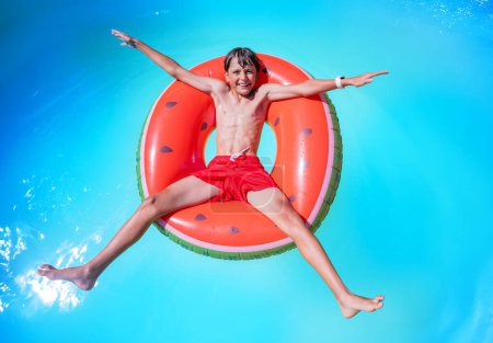 Foto de Momento despreocupado como un joven nadador adolescente en traje de baño rojo festivo se relaja en una rebanada de sandía flotar en una piscina de agua serena - Imagen libre de derechos