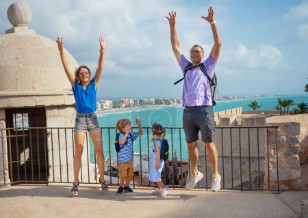 Téléchargez les photos : Jeune famille voyageant avec des enfants sauter heureux sur le point de vue, Peniscola ville et le littoral en arrière-plan, Espagne - en image libre de droit