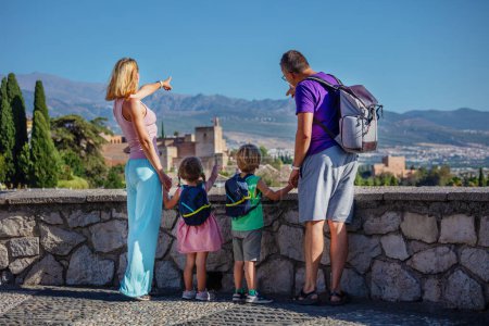 Téléchargez les photos : Jeunes touristes avec des enfants debout sur le point de vue, point à l'ancienne citadelle célèbre Alhambra sur leur tournée touristique d'été à Grenade, Espagne - en image libre de droit