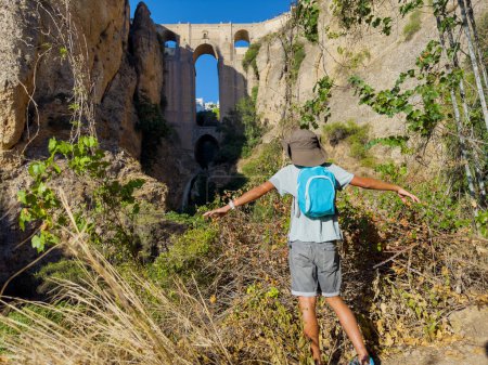 Téléchargez les photos : Regard touristique heureux sur le magnifique Puente Nuevo à Ronda depuis le fond de la gorge d'El Tajo lors de sa visite touristique en Espagne - en image libre de droit