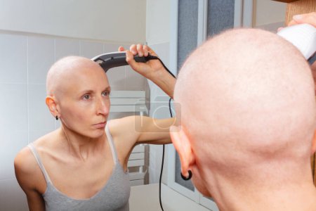 Foto de Mujer fuerte y segura que lucha contra la pérdida de cabello después del tratamiento del cáncer afeitándose la cabeza con un zumbador, concentrándose en la imagen del espejo - Imagen libre de derechos