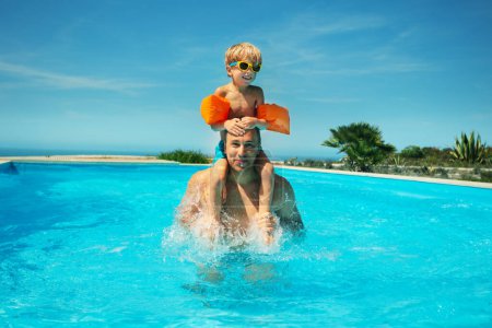 Photo for Young child in orange arm floaties and sunglasses rides on the shoulders of an adult male in a sunlit swimming pool - Royalty Free Image