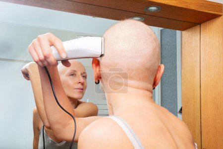 Foto de Fuerte lucha de la mujer con el cáncer es auto-cuidado utilizando una máquina de afeitar eléctrica de mano para afeitarse la cabeza delante de un espejo de baño - Imagen libre de derechos