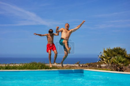 Foto de Dos individuos, padre e hijo saltan alegremente a una piscina en un día soleado con un cielo azul claro, mar al fondo - Imagen libre de derechos