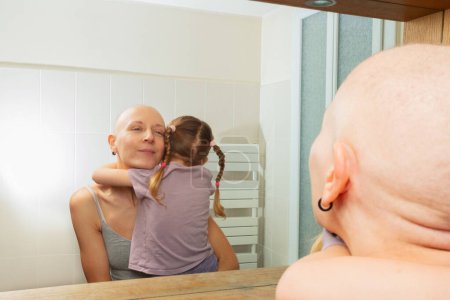 Foto de Una mujer calva luchando contra el cáncer abrazando a una joven, reflejando amor y apoyo, como se ve en el reflejo del espejo - Imagen libre de derechos
