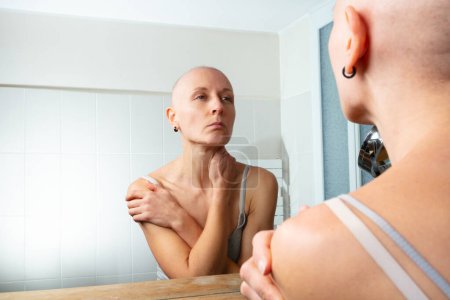 Téléchargez les photos : Regardant dans le miroir de la salle de bain, une femme à la tête chauve tient délicatement son cou et son épaule, perdue dans la pensée - en image libre de droit