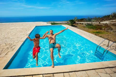 Foto de Un par de alegres nadadores, hijo y padre dan un salto hacia atrás en una refrescante piscina con una impresionante vista al mar en un día brillante - Imagen libre de derechos